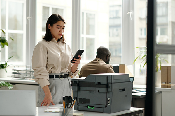 Sicheres Drucken: Frau steht mit Smartphone in der Hand vor einem Drucker
